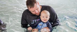 Young boy baby smiles at camera while Dad in black swim shirt holds him in the water